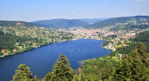 Vue panoramique du lac de Gérardmer, avec des voiliers sur l'eau bleue, entouré de collines verdoyantes et d'une ville pittoresque.