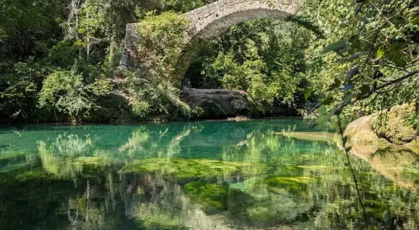 Pont de pierre envahi par la végétation au-dessus des eaux turquoise des gorges de la Siagne, reflétant le ciel et la végétation luxuriante.
