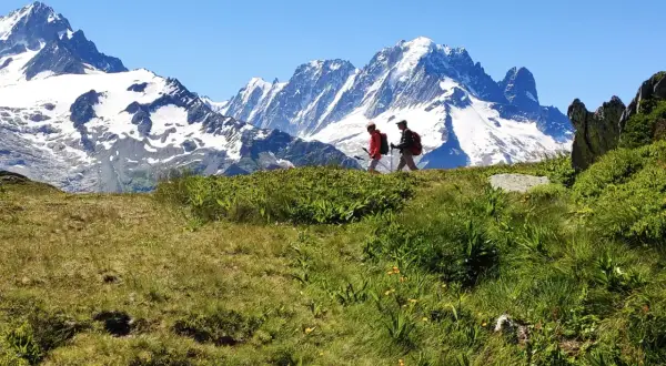Deux randonneurs marchent sur une crête herbeuse face à des montagnes enneigées.
