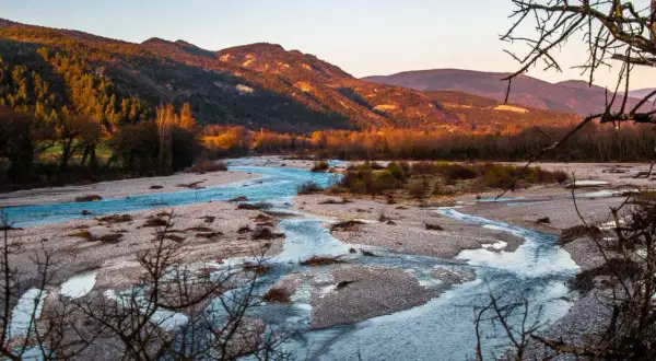Le **saut de la Drôme** offre un paysage de rivière aux eaux bleues vives serpentant entre des bancs de galets, sous des montagnes dorées par le soleil couchant.