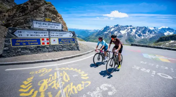 Deux cyclistes gravissent le col du Galibier, un paysage alpin spectaculaire sous un ciel bleu éclatant.