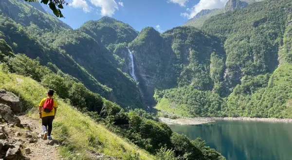 Randonneur admirant la cascade et le lac de Toulouse, entourés de montagnes verdoyantes sous un ciel bleu.