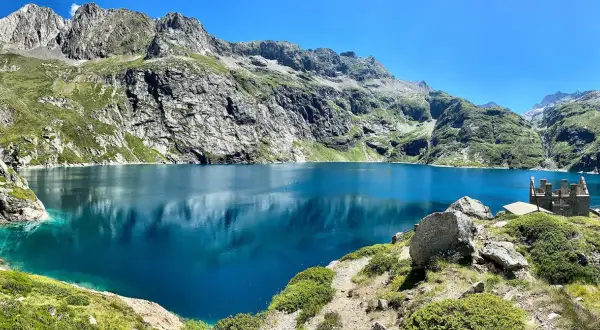 Le lac de Caillauas, d'un bleu profond, reflète les montagnes escarpées et verdoyantes sous un ciel clair. Une structure rocheuse rappelle un château sur la rive.