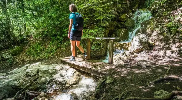 Une randonneuse traverse un pont en bois près de la cascade de Morette, entourée d'une végétation luxuriante.