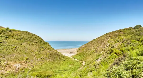 Vue sur la mer à travers la vallée du Lude, bordée de collines verdoyantes et d'une plage de sable.