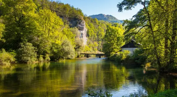 Rivière paisible reflétant la verdure luxuriante, un pont discret et une maisonnette au bord de l'eau, le tout sous un ciel bleu clair, évoquant la sérénité de la cascade de Syratus.