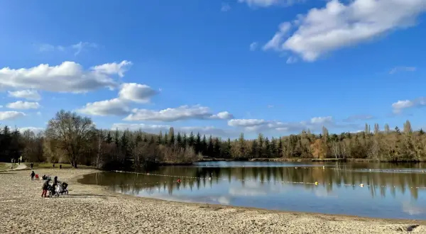 Plage de sable fin au bord d'un lac paisible, avec des familles profitant du soleil. La **cascade** de la **forêt de Fontainebleau** se reflète dans le **lac** sous un ciel bleu parsemé de nuages.