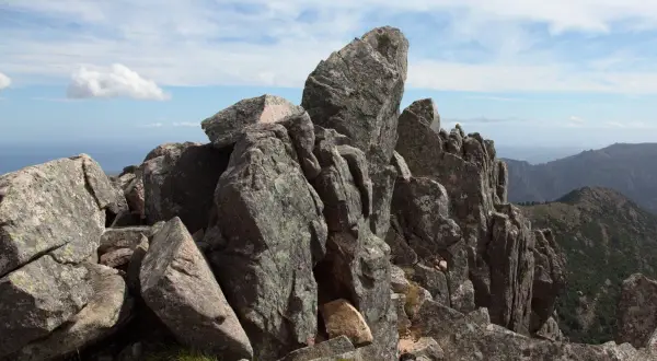 Rochers escarpés au sommet de la Punta di a vacca morta, avec des montagnes en arrière-plan sous un ciel bleu et nuageux.