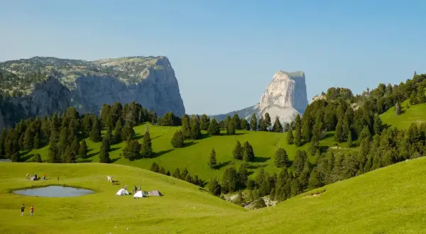 Un campement de tentes et de randonneurs sur une prairie verdoyante, avec des montagnes imposantes en arrière-plan. Le **pas de l'aiguille** est visible au loin.