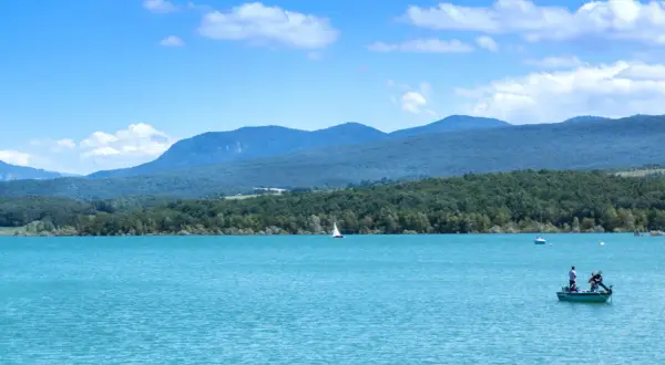 Le lac de Montbel, aux eaux turquoise, accueille un voilier et un petit bateau de pêche sous un ciel bleu parsemé de nuages.