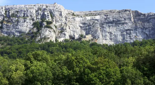 Vue du pic de Bertagne, une falaise imposante surplombant une forêt verdoyante. Un petit bâtiment est visible sur le flanc de la falaise.