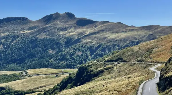 Route sinueuse dans les montagnes, avec des pâturages dorés et une forêt dense. Pas de Peyrol, un paysage grandiose sous un ciel bleu.
