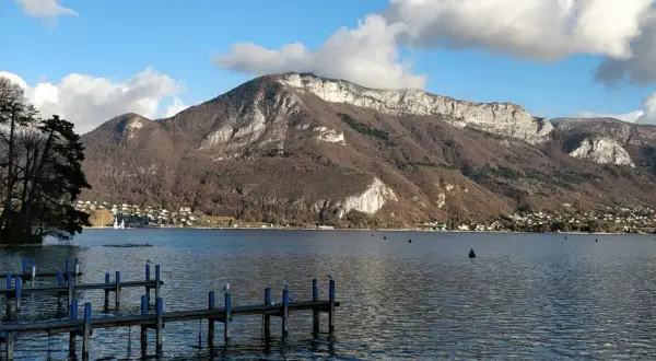Vue de la webcam du lac d'Annecy, avec un ponton en bois et des montagnes majestueuses en arrière-plan.