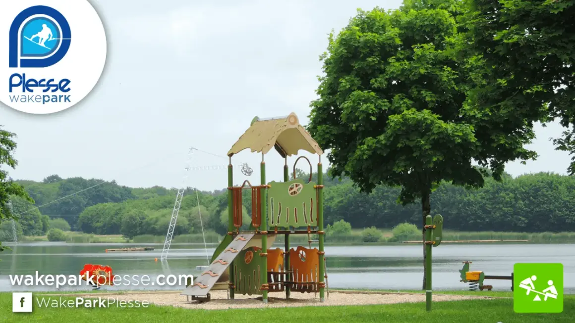 Aire de jeux près de l'étang de Buhel, avec un toboggan et un portique. Le logo WakePark Plessé est visible.