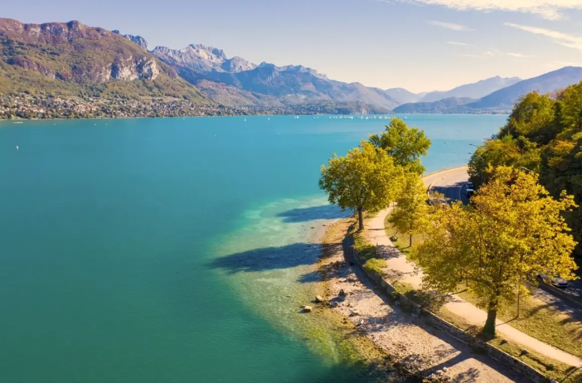 Vue aérienne du lac d'Annecy, ses eaux turquoise bordées par une route sinueuse et des arbres aux couleurs d'automne. La taille du lac est impressionnante.