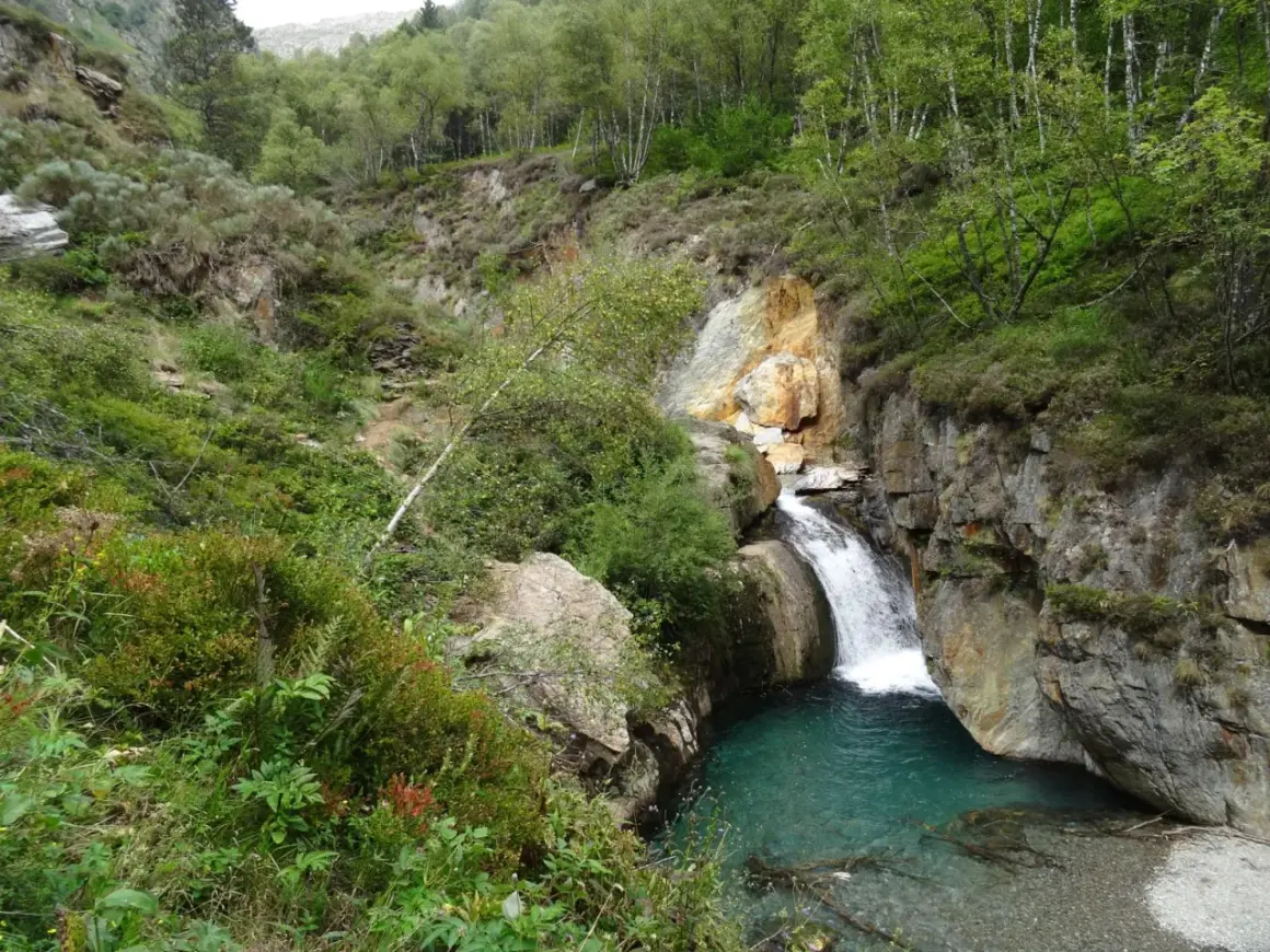 La cascade de l'Artigue dévale dans un bassin d'eau turquoise, entourée de roches et d'une végétation luxuriante.