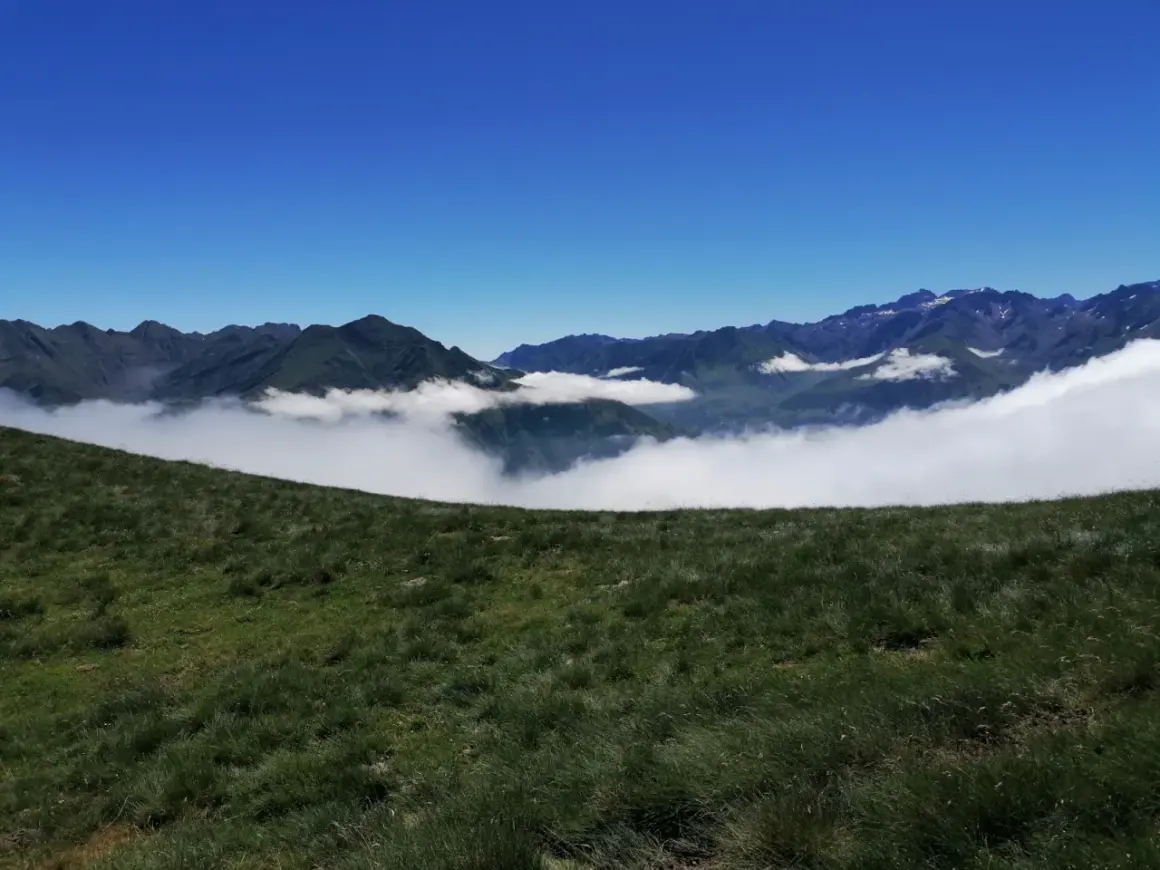 Vue panoramique depuis le col de Riou, avec des montagnes verdoyantes émergeant d'une mer de nuages sous un ciel bleu éclatant.