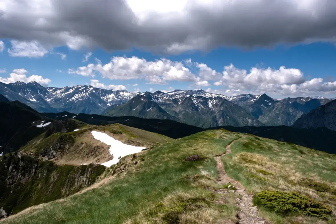 Sentier rocailleux menant vers le pic de Tarbesou, avec des montagnes enneigées en arrière-plan sous un ciel nuageux.