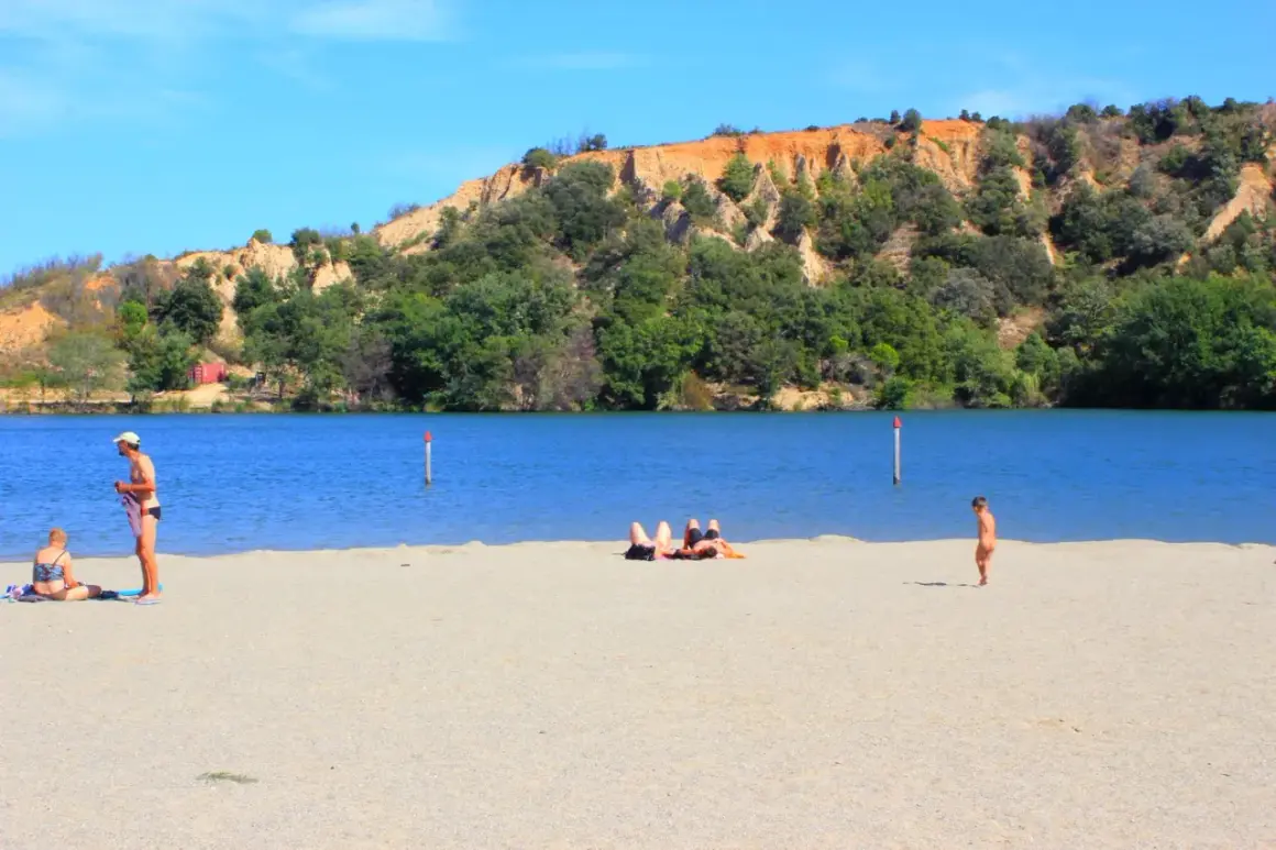 Plage de sable fin au lac de Vinça, avec des baigneurs profitant du soleil et de la vue sur la colline verdoyante.
