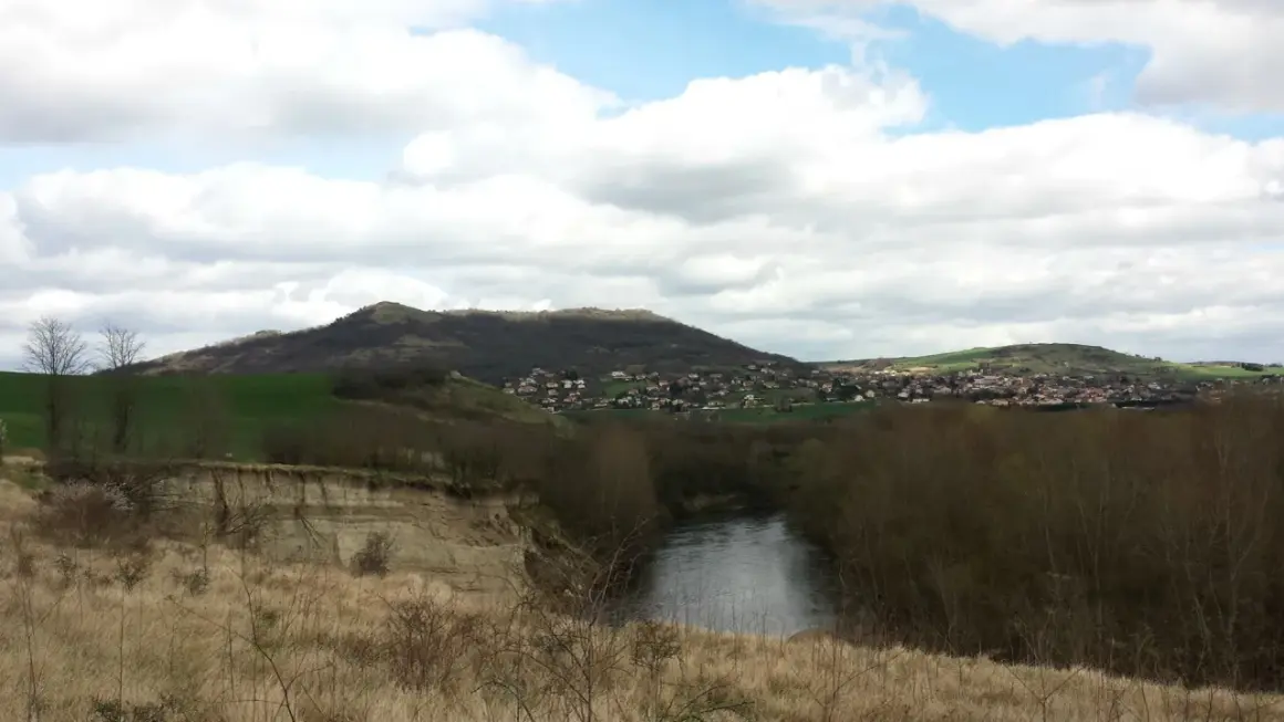 Vue panoramique d'une rivière sinueuse bordée d'arbres, avec une falaise de malmouche en contrebas et un village niché sur une colline verdoyante.