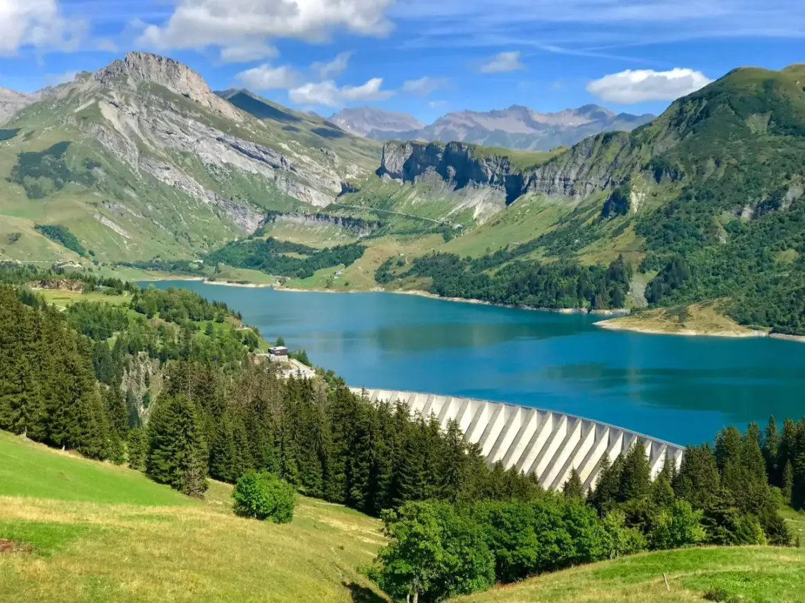 Vue panoramique du lac de Roselend, un barrage imposant bordé de forêts verdoyantes et de montagnes majestueuses sous un ciel bleu éclatant.