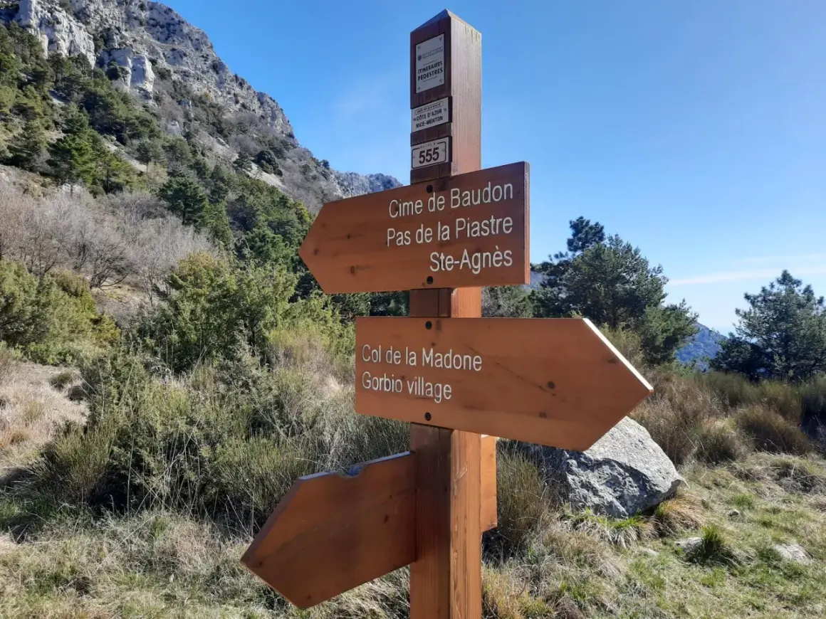 Panneau indicateur de randonnée : direction Cime de Baudon, Pas de la Piastre, Ste-Agnès, Col de la Madone et Gorbio village. Paysage montagneux.