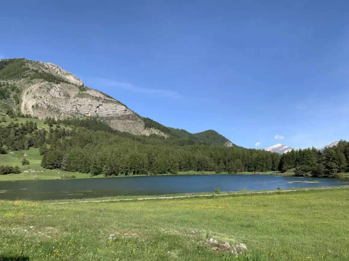 Le lac de Barbeyroux, paisible, reflète le ciel bleu. Des montagnes rocheuses et une forêt dense bordent ses rives verdoyantes.