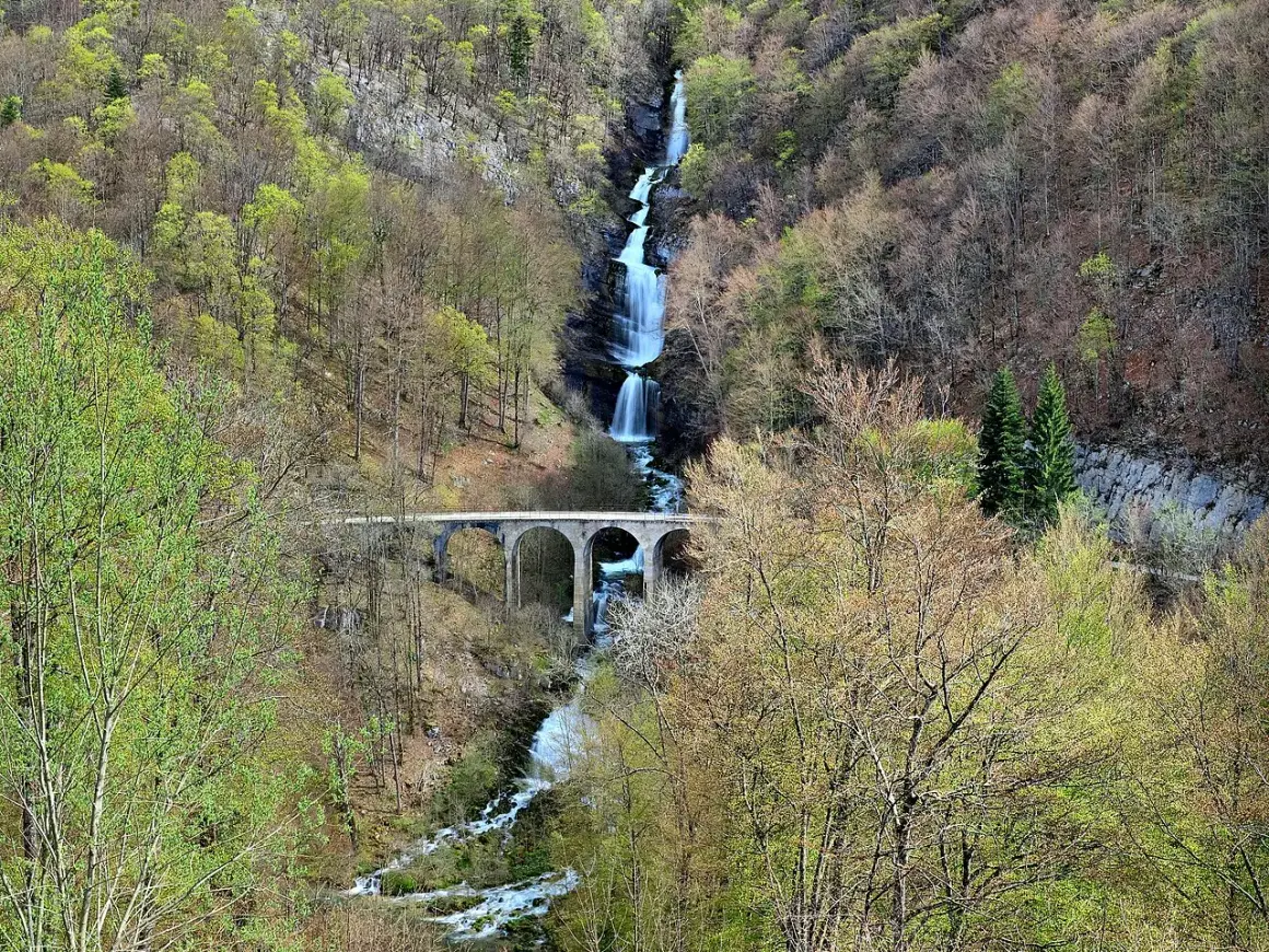 La cascade du Bief de la Ruine dévale une falaise rocheuse, traversée par un pont en arc. La nature verdoyante environnante annonce le printemps.