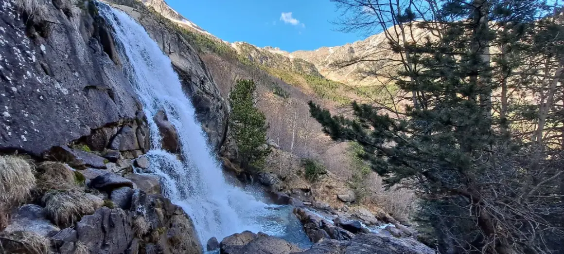 La cascade des Molières dévale une paroi rocheuse, entourée de pins et de montagnes enneigées sous un ciel bleu.