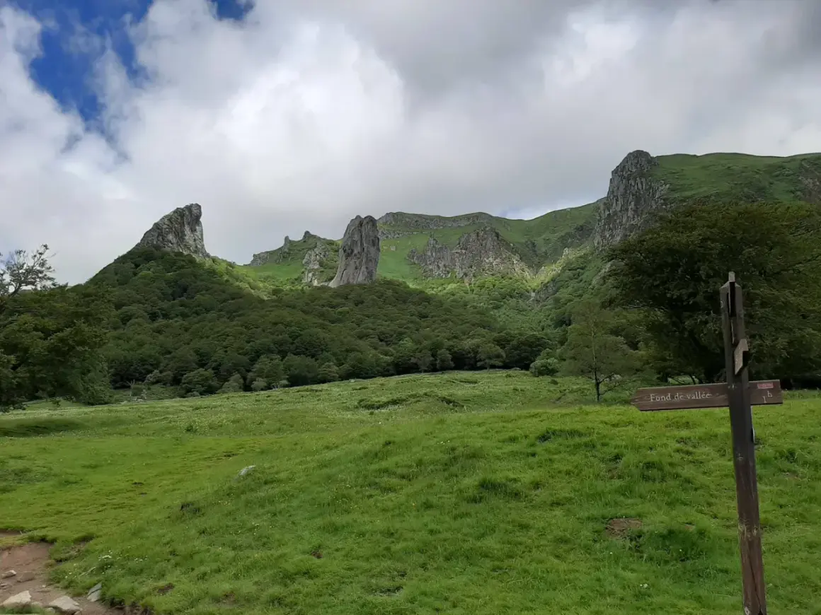 Paysage montagneux verdoyant avec des formations rocheuses spectaculaires. Un panneau indique "Fond de vallée", peut-être le chemin vers la cascade de la biche.