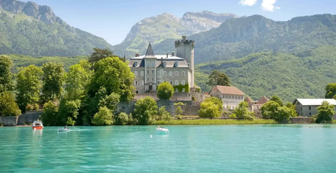 Un château majestueux surplombe un lac turquoise, invitant à aller à Annecy. Des montagnes verdoyantes en arrière-plan.