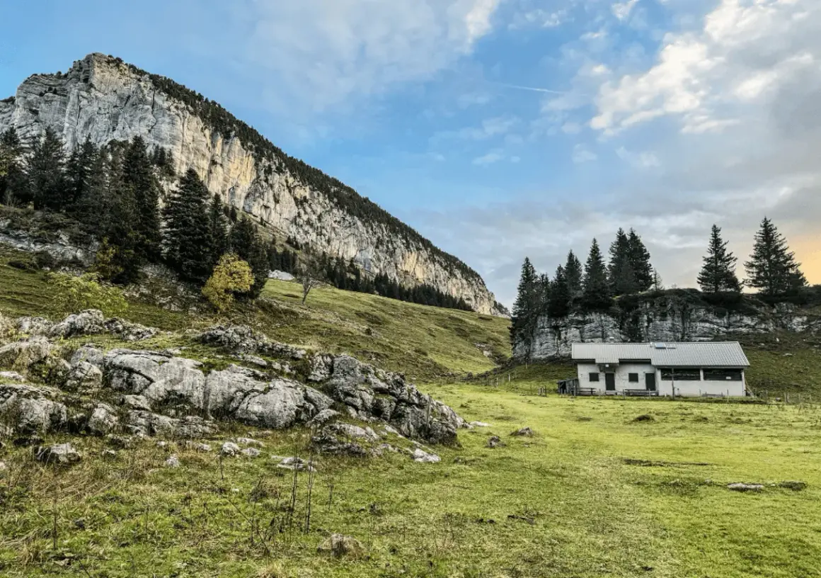 Un chalet blanc se dresse dans un pré verdoyant au pied d'une falaise imposante. Le col de l'Alpette offre un paysage alpin paisible.