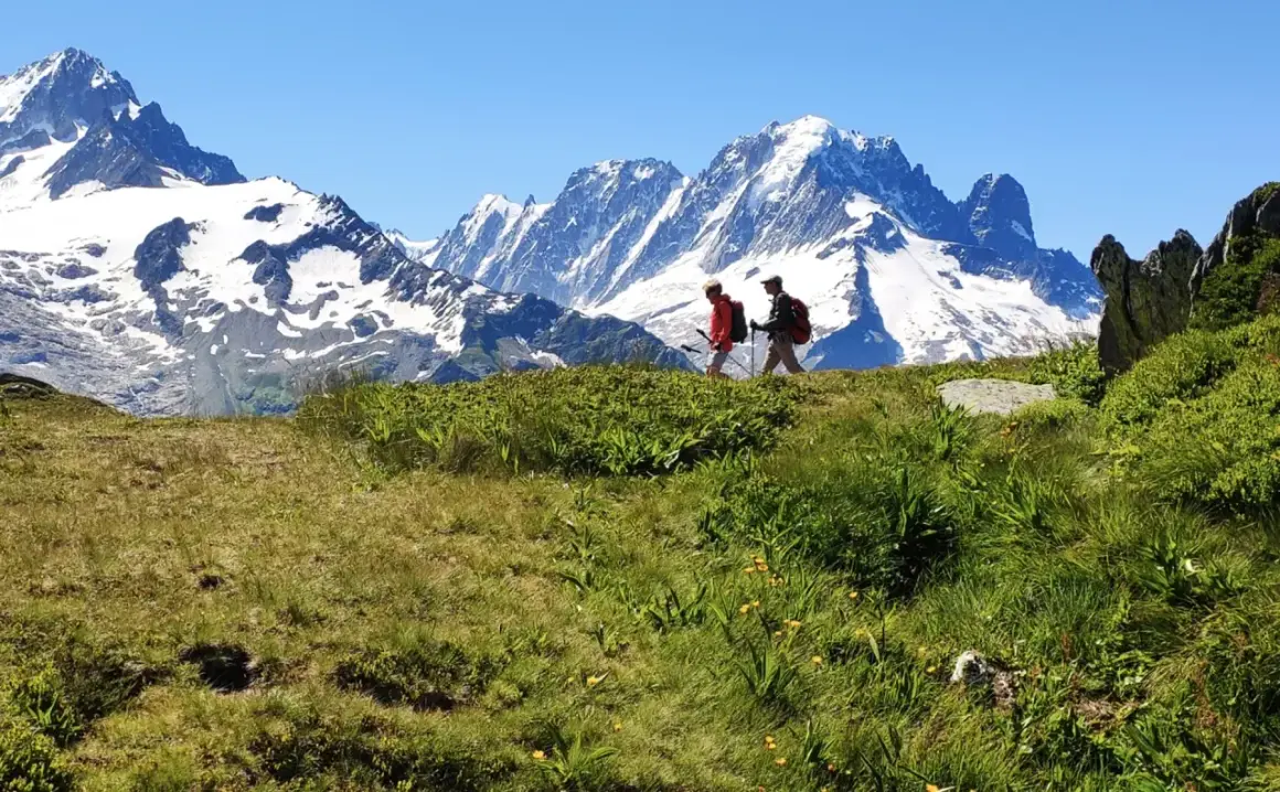 Deux randonneurs marchent sur une crête herbeuse face à des montagnes enneigées.