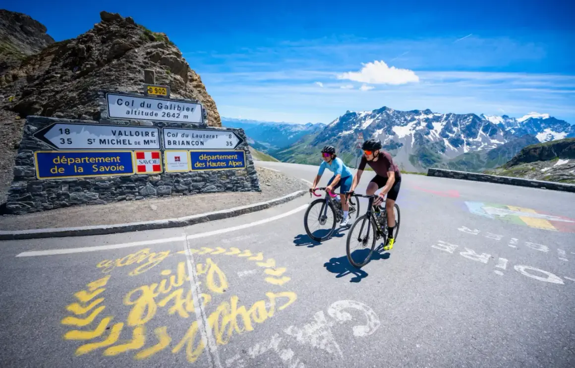 Deux cyclistes gravissent le col du Galibier, un paysage alpin spectaculaire sous un ciel bleu éclatant.