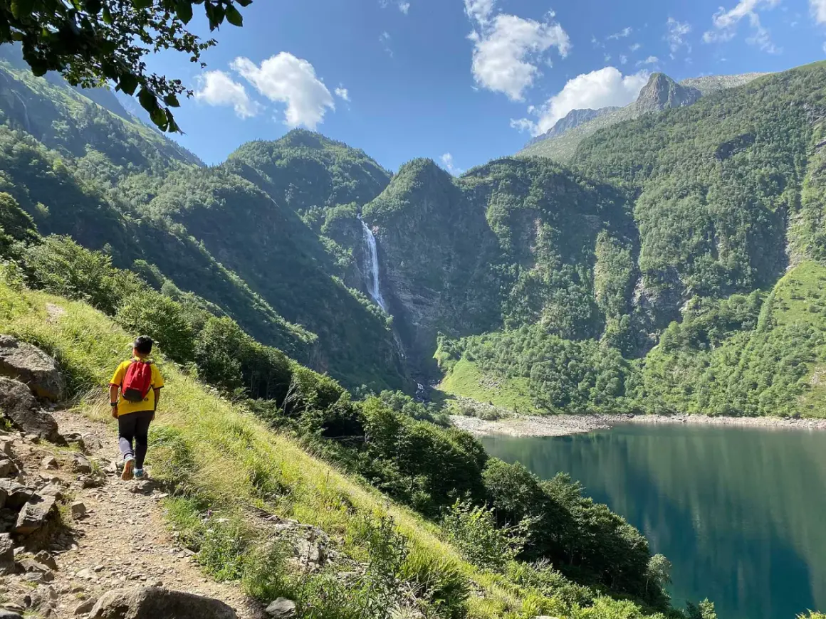 Randonneur admirant la cascade et le lac de Toulouse, entourés de montagnes verdoyantes sous un ciel bleu.