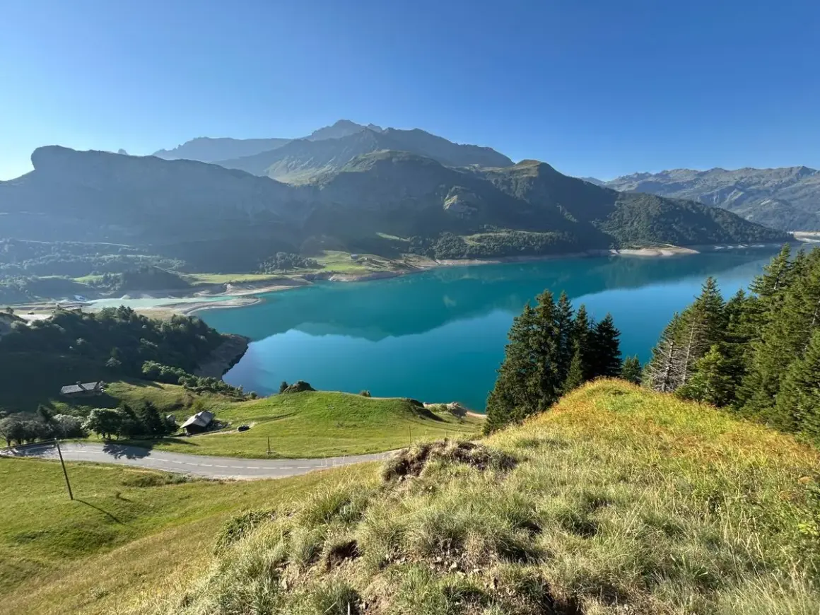 Vue panoramique du lac de la Gittaz aux eaux turquoise, entouré de montagnes verdoyantes et d'une route sinueuse.