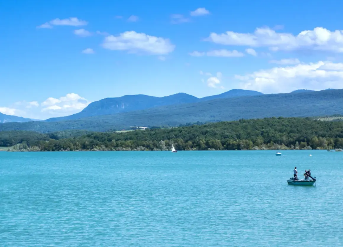 Le lac de Montbel, aux eaux turquoise, accueille un voilier et un petit bateau de pêche sous un ciel bleu parsemé de nuages.