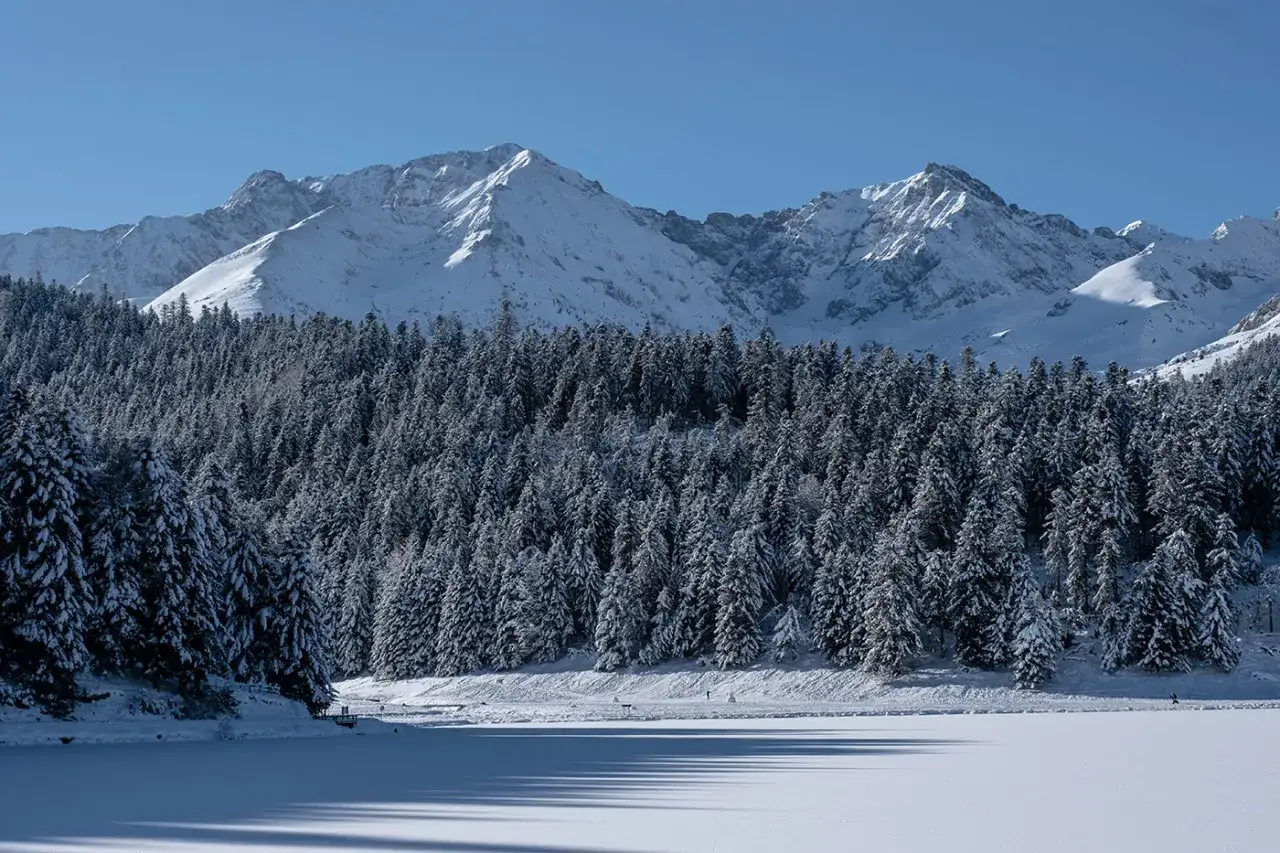Paysage hivernal du lac de Payolle, avec des montagnes enneigées en arrière-plan et une forêt de sapins recouverts de neige.