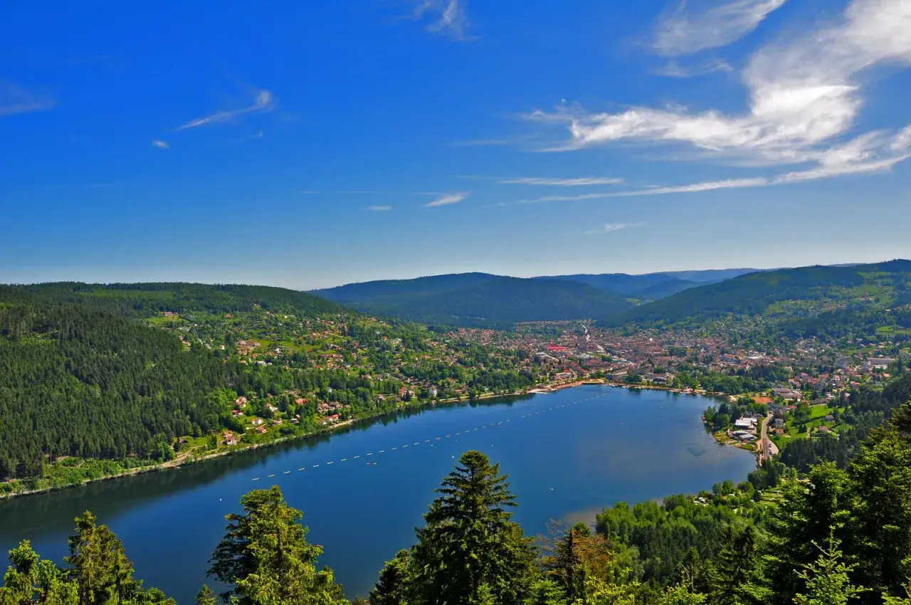 Vue panoramique du lac de G&eacute;rardmer, entour&eacute; de for&ecirc;ts verdoyantes et de maisons pittoresques, sous un ciel bleu &eacute;clatant.