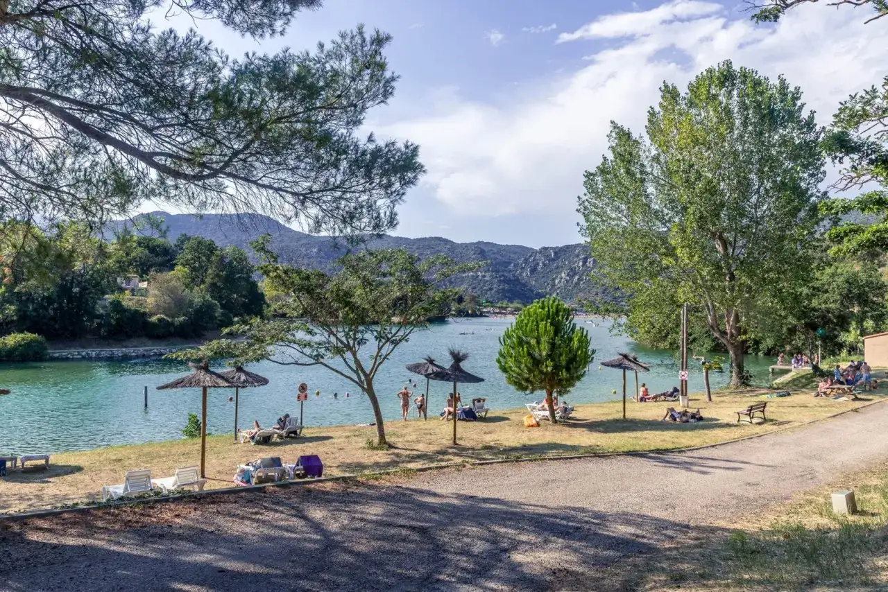 Bord du lac de Vinça, des parasols et des transats invitent à la détente. Des baigneurs profitent de l'eau turquoise sous un ciel bleu.