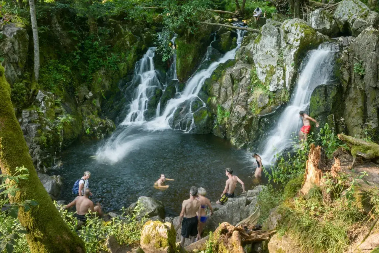 Des baigneurs profitent d'un saut du bouchot dans une cascade rocheuse entourée de verdure.