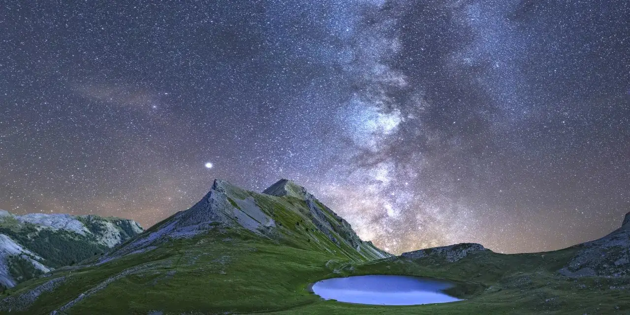 Le lac des Pises sous une voie lact&eacute;e spectaculaire. Montagnes verdoyantes et ciel &eacute;toil&eacute; se refl&egrave;tent dans l'eau paisible.