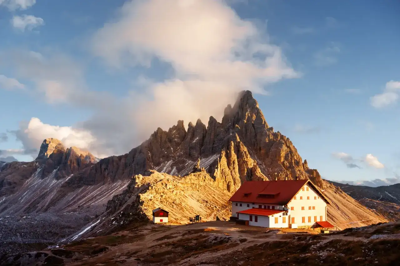 Refuge sur l'Alta Via 2, au pied de montagnes spectaculaires baign&eacute;es par le soleil du matin. Nuages et pics rocheux se m&ecirc;lent dans un paysage alpin grandiose.