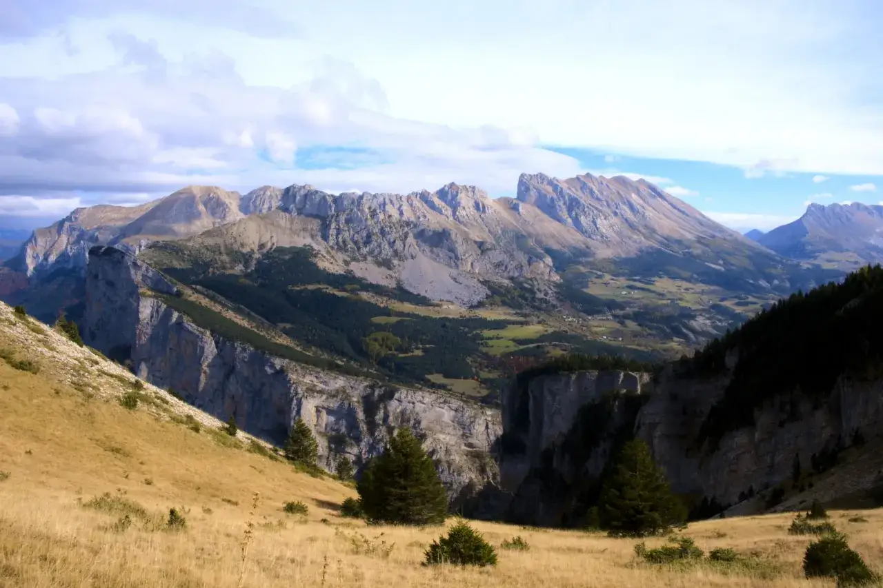 Vue panoramique sur le col du Festre, avec ses falaises abruptes, ses for&ecirc;ts de pins et ses prairies dor&eacute;es sous un ciel nuageux.