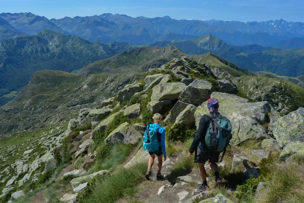 Deux randonneurs escaladent un sentier rocheux vers le pic des 3 Seigneurs, offrant une vue panoramique sur les Pyr&eacute;n&eacute;es verdoyantes.