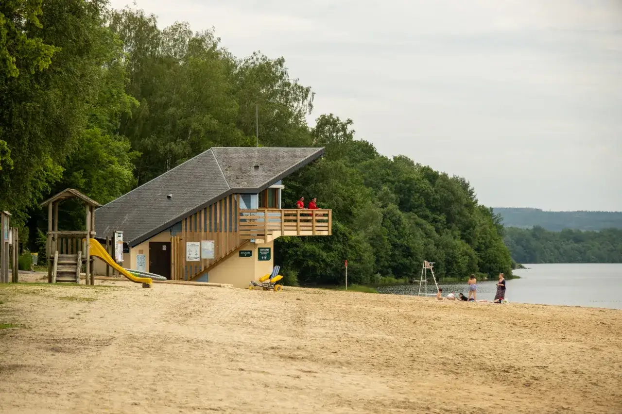 Plage de sable fin au lac des vieilles forges, avec un bâtiment de secours, une aire de jeux et des baigneurs profitant de la journée.