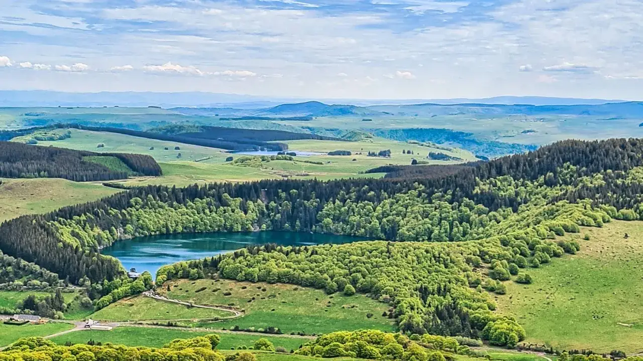 Vue panoramique du Puy de Chambourguet, avec son lac entour&eacute; de for&ecirc;ts verdoyantes et de collines ondulantes sous un ciel bleu.