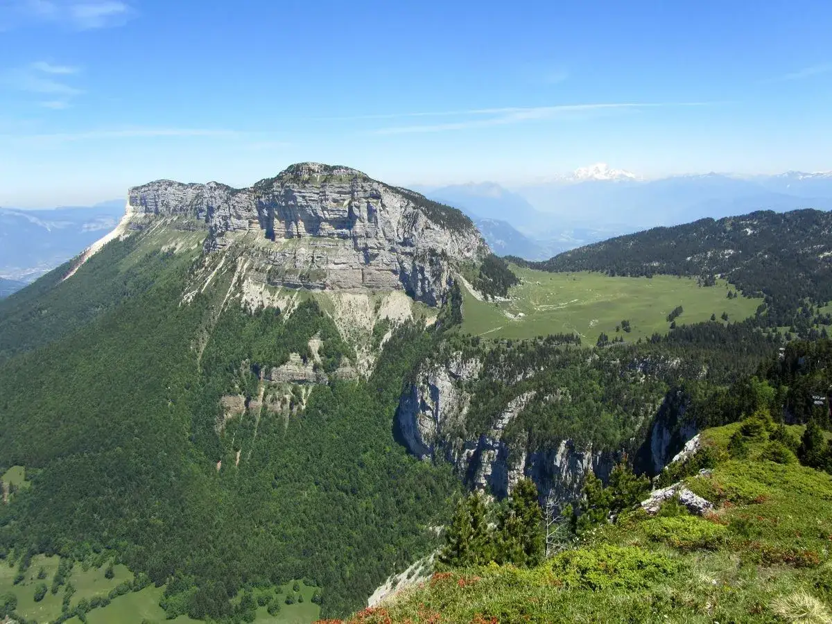 Vue panoramique depuis le col de l'Alpette, avec ses falaises imposantes, ses forêts verdoyantes et les sommets enneigés en arrière-plan.