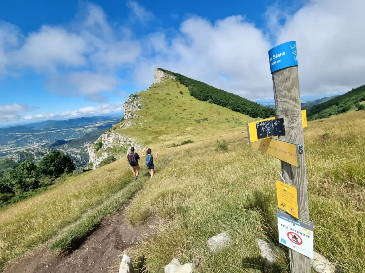 Deux randonneurs sur un sentier herbeux, dominant un paysage de montagnes. Un panneau indique