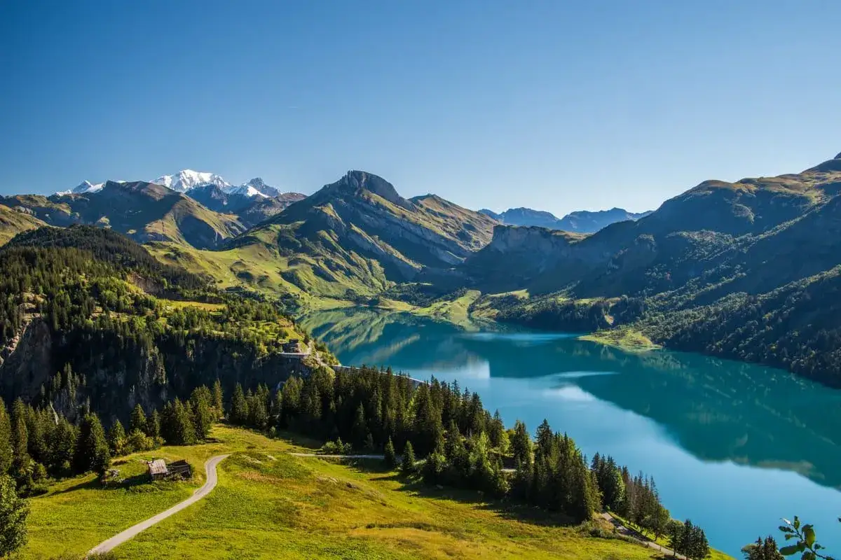 Vue panoramique du lac de Roselend, aux eaux turquoise, entouré de montagnes verdoyantes et de forêts de pins sous un ciel bleu clair.